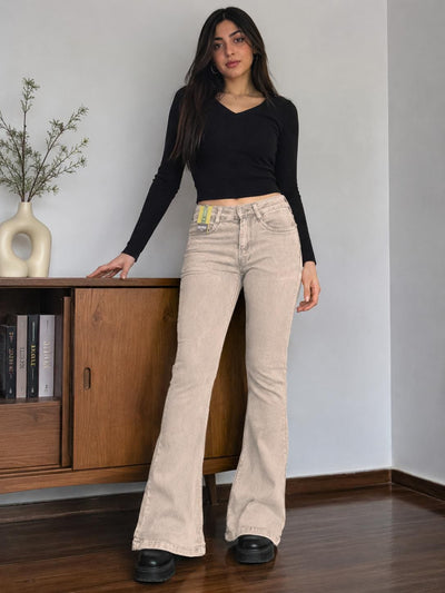 A woman stands indoors wearing a fitted black long-sleeve crop top and light beige flared jeans with black platform shoes. She is posed in front of a wooden cabinet against a plain light gray wall.